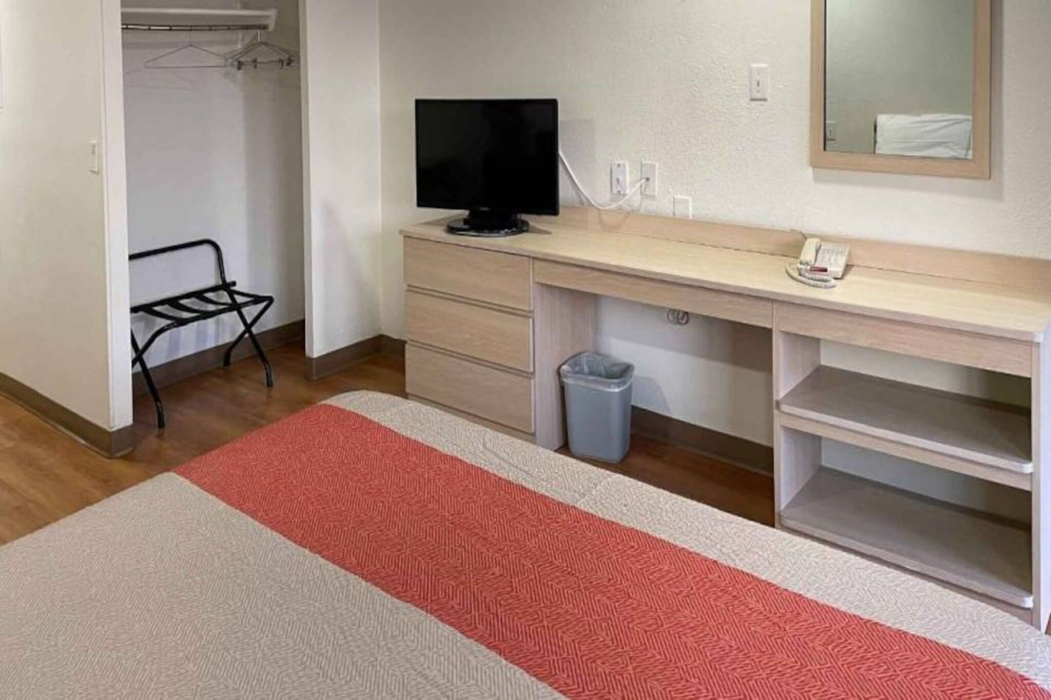 A hotel room with a desk, a television, a telephone, a trash bin, and a closet with a hanging rack.