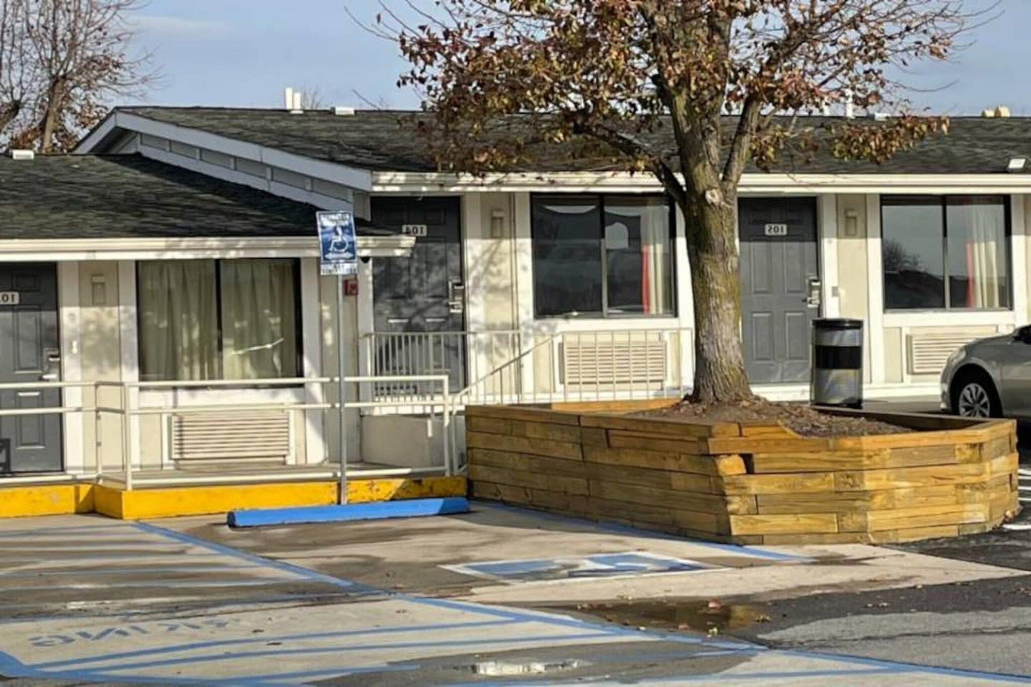 Un edificio con acceso mediante rampa, un árbol en una jardinera y un aparcamiento con plazas delimitadas.