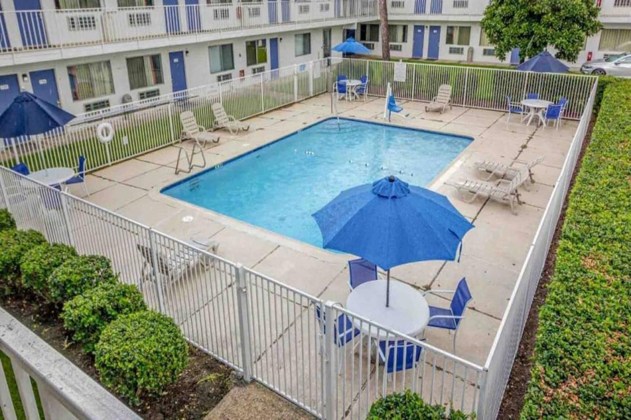 A swimming pool with lounge chairs and umbrellas, surrounded by a fence, in a courtyard with a building in the background.