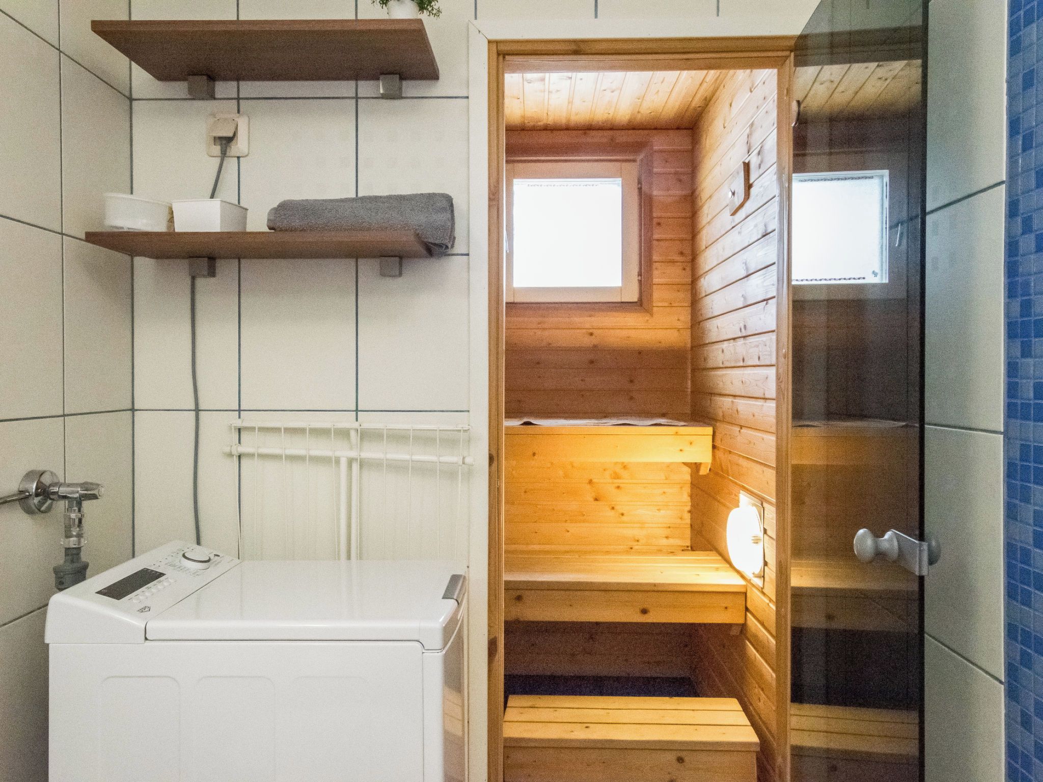 A small, enclosed sauna with wooden walls and a window, a white washing machine, and a tiled wall with a towel rack.