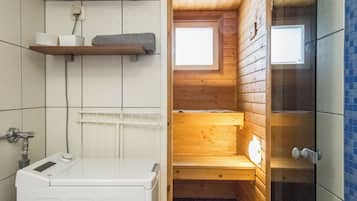 Uma sauna pequena e fechada, com paredes de madeira e uma janela, uma máquina de lavar branca e um revestimento de azulejos com um porta-toalhas.