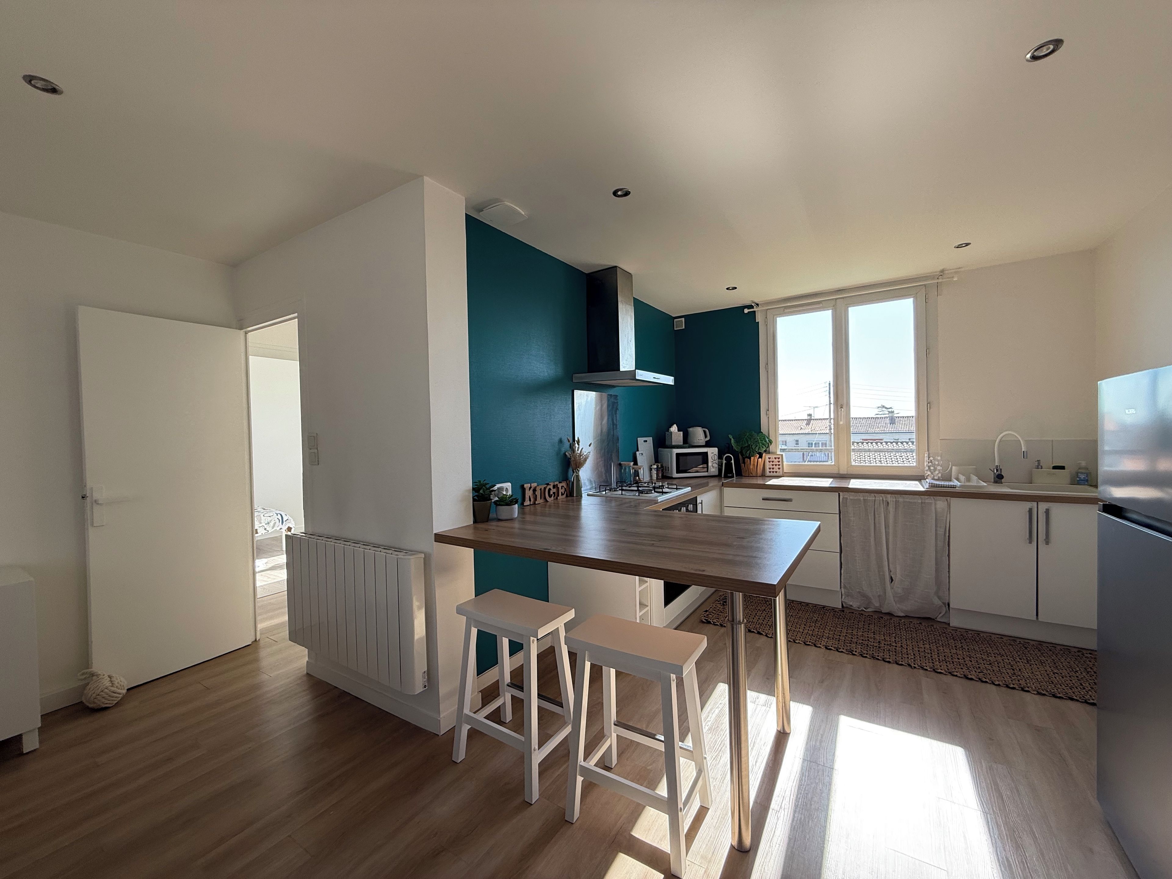 Una cocina moderna con encimera de madera, taburetes blancos, microondas integrado y una ventana que deja entrar luz natural.