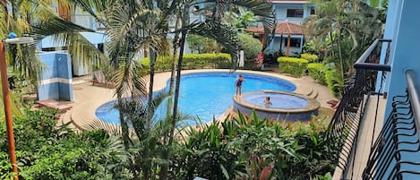 Une piscine entourée d’une végétation luxuriante et un bâtiment bleu avec un toit recouvert de tuiles.