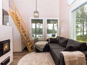A cozy cabin interior with a wooden staircase, a fireplace, a sofa, and large windows offering a view of the outdoors.