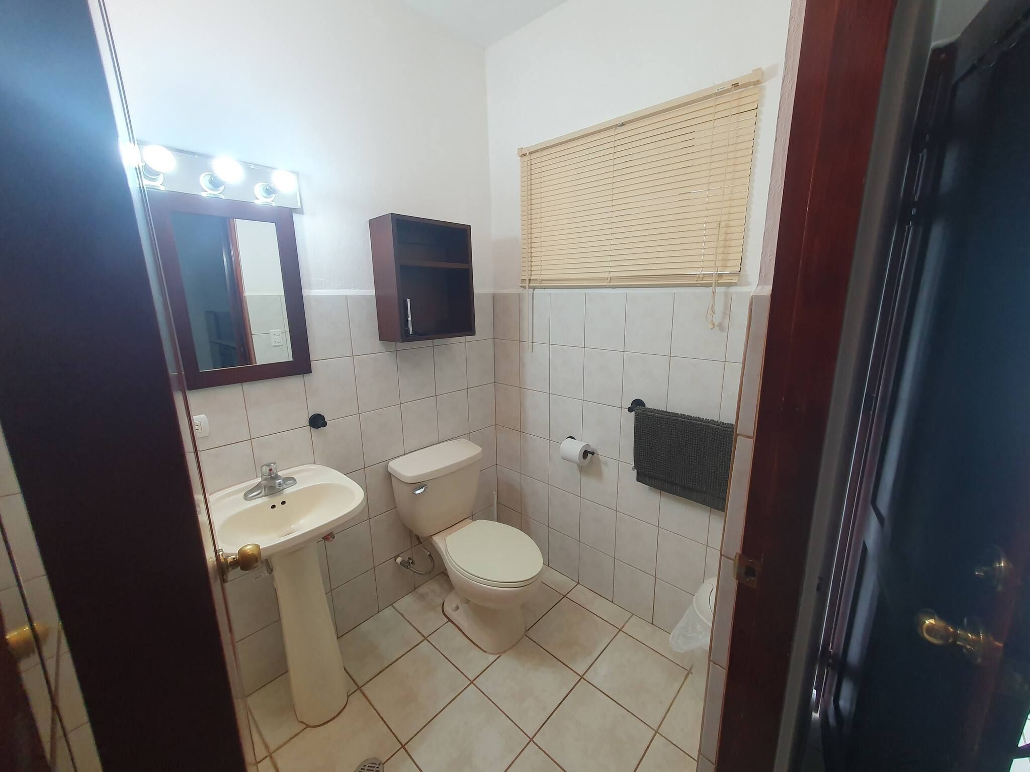 A bathroom with a sink, toilet, mirror, and a window with blinds.