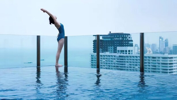 Una persona in costume blu che esegue una posa yoga su una piscina a sfioro con fondo trasparente, con una skyline cittadina sullo sfondo.