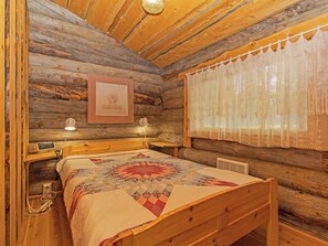 Une chambre chaleureuse dans un chalet en rondins, avec un lit en bois, une courtepointe, un tableau encadré au mur et un rideau en dentelle devant la fenêtre.