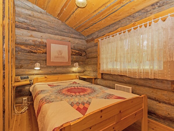 Une chambre chaleureuse dans un chalet en rondins, avec un lit en bois, une courtepointe, un tableau encadré au mur et un rideau en dentelle devant la fenêtre.