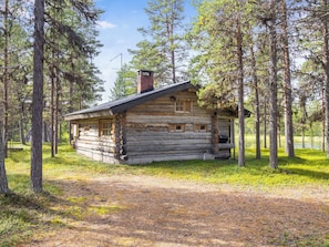 Une cabane rustique en rondins de bois, dotée d’une cheminée, entourée de grands arbres et d’une zone herbeuse.