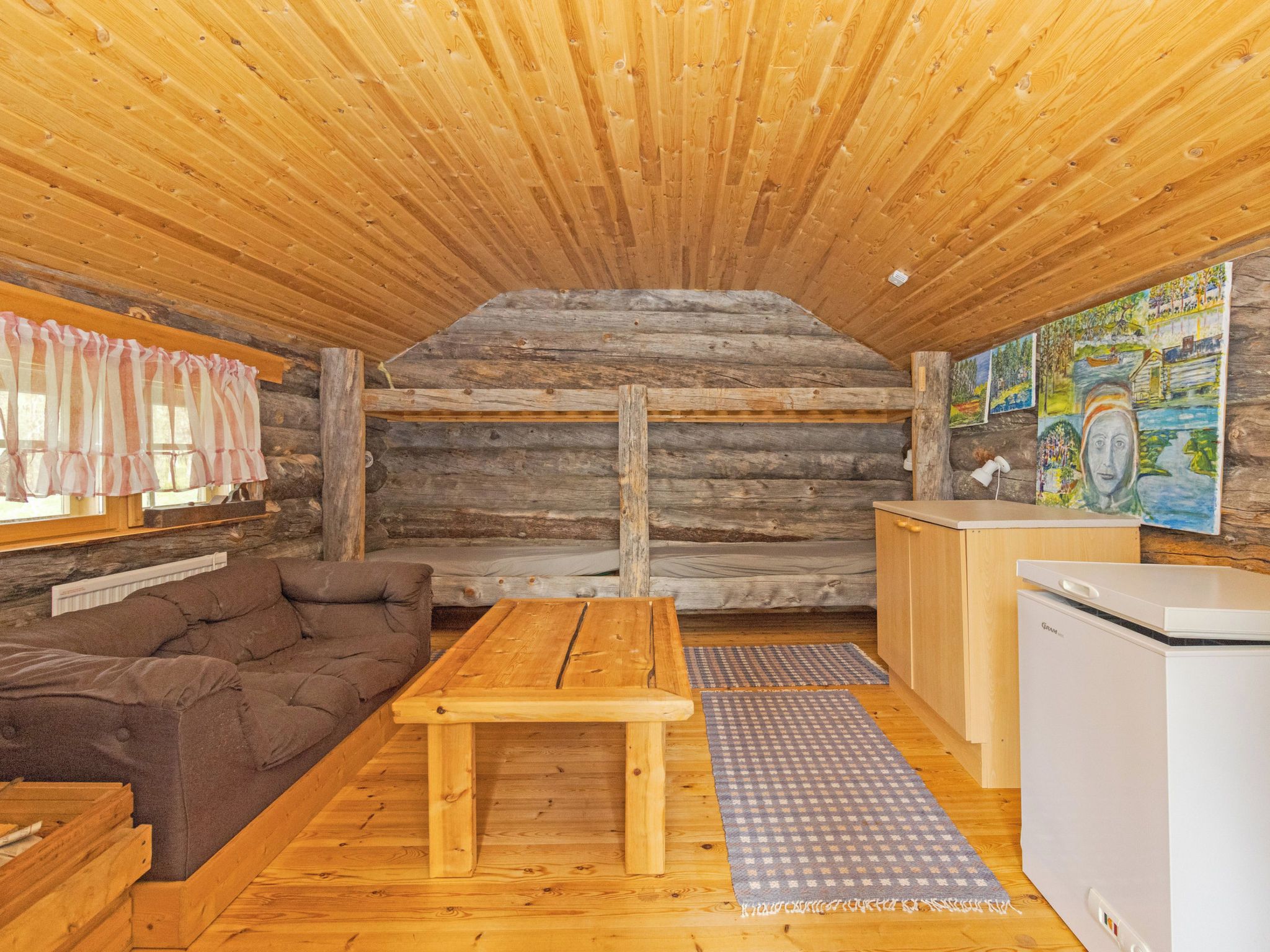Interior acolhedor de uma cabana, com teto de madeira, um sofá, uma mesa de madeira, geladeira e obras de arte nas paredes.