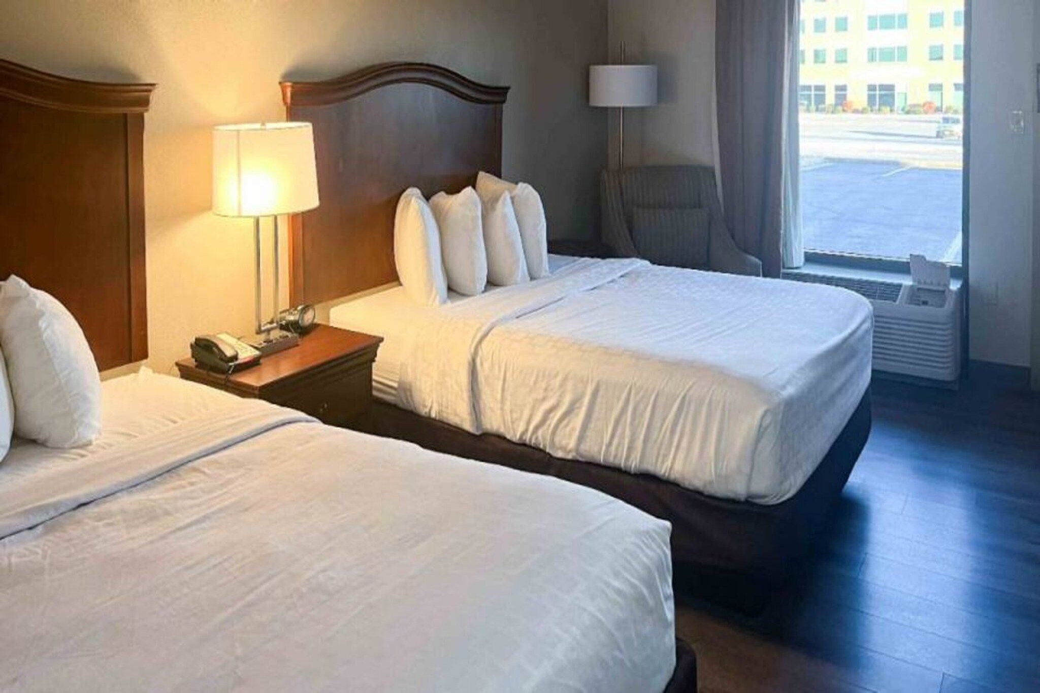 A hotel room with two beds, a nightstand with a lamp, and a view of the outside.