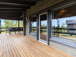 Ein überdachter Außenbereich mit Holzterrasse, einem Tisch mit Stühlen und großen Fenstern mit Blick auf Bäume und ein Gebäude.