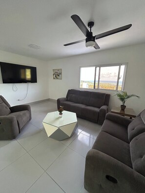 A modern living room with a flat-screen TV, a ceiling fan, a sofa, a coffee table, and a window with a view.