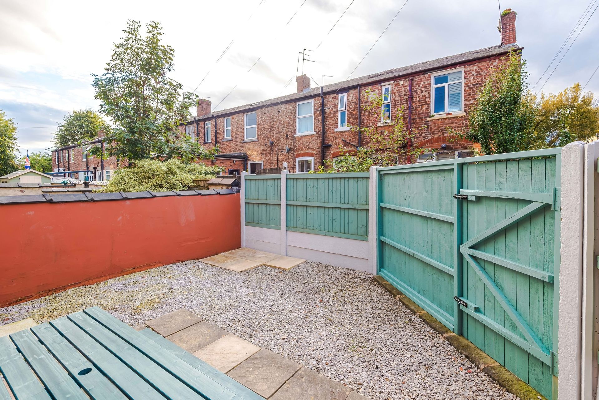 A residential backyard with a red wall, a green gate, and a gravel path leading to a wooden bench.