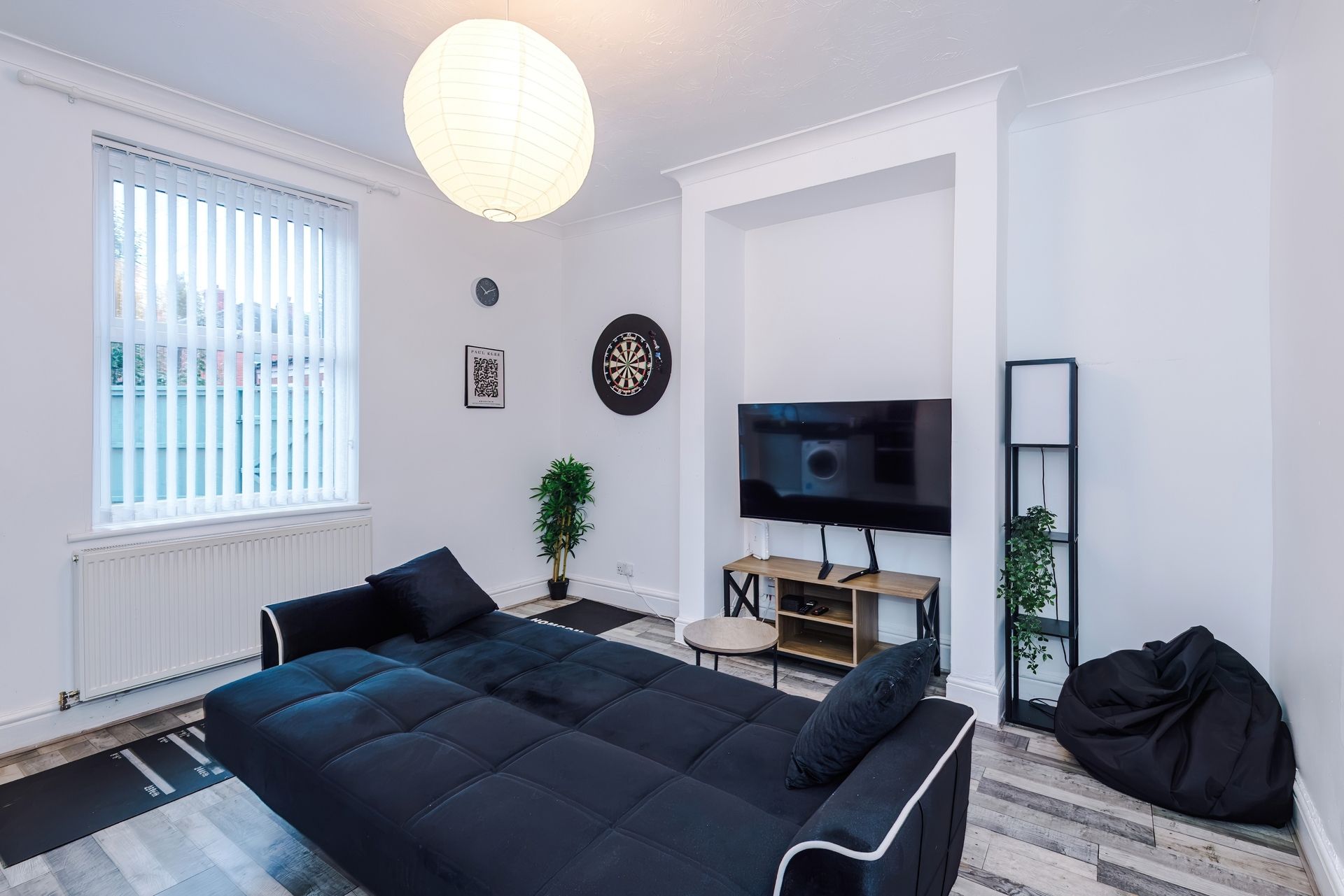 A modern living room with a large black sofa, a flat-screen TV, a dartboard, and a potted plant.