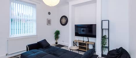 A modern living room with a large black sofa, a flat-screen TV, a dartboard, and a potted plant.