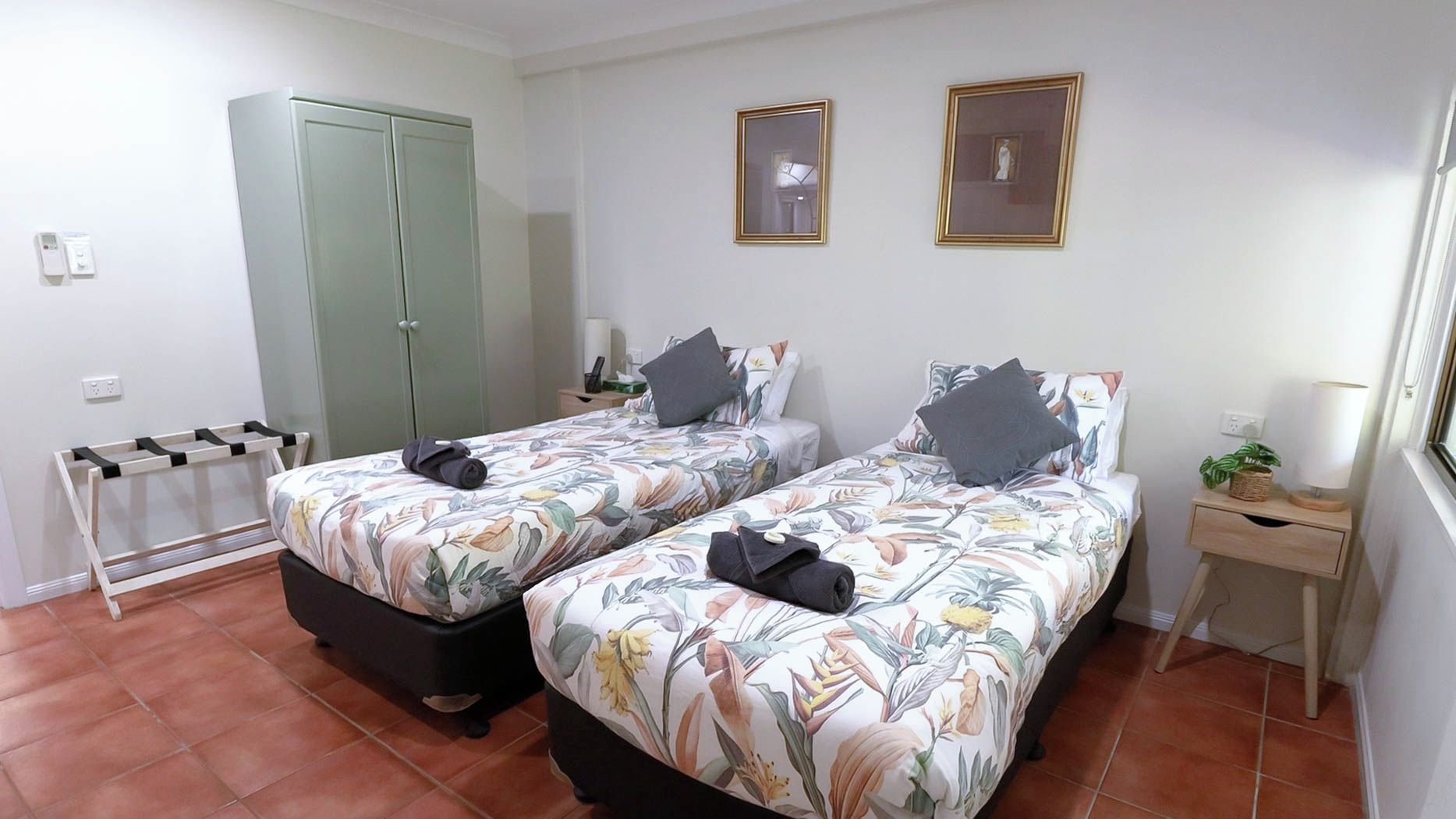 A room with two beds, a wardrobe, a bedside table with a lamp, and a small window.