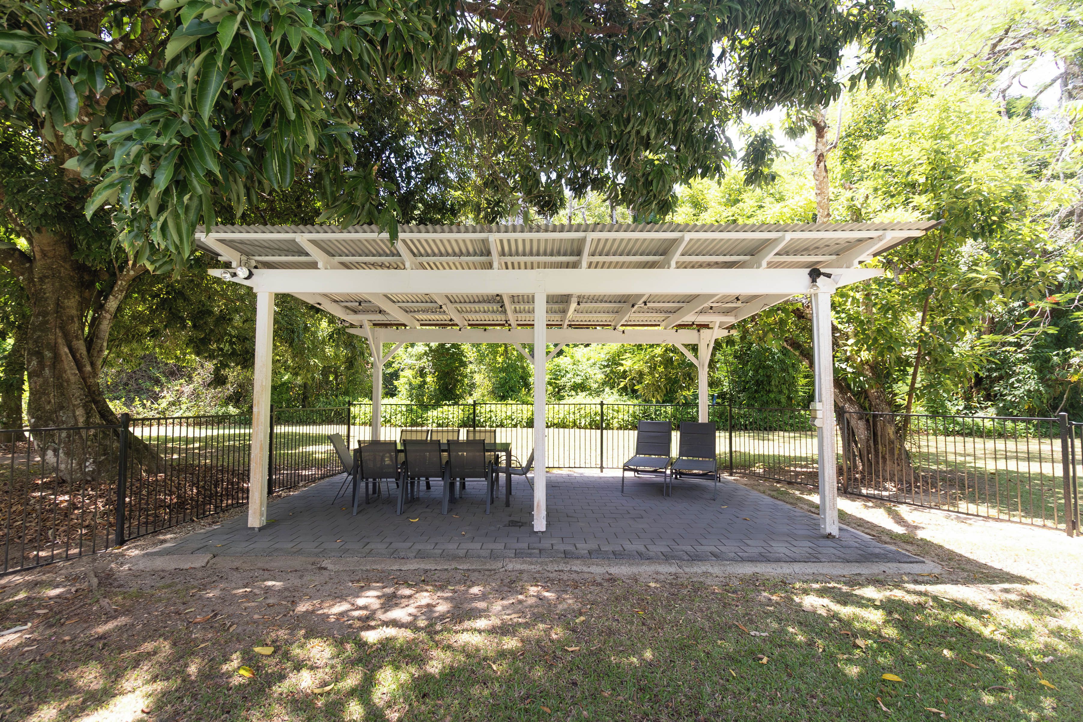 Un espace extérieur abrité, doté de tables et de chaises, entouré d’arbres et d’une clôture.