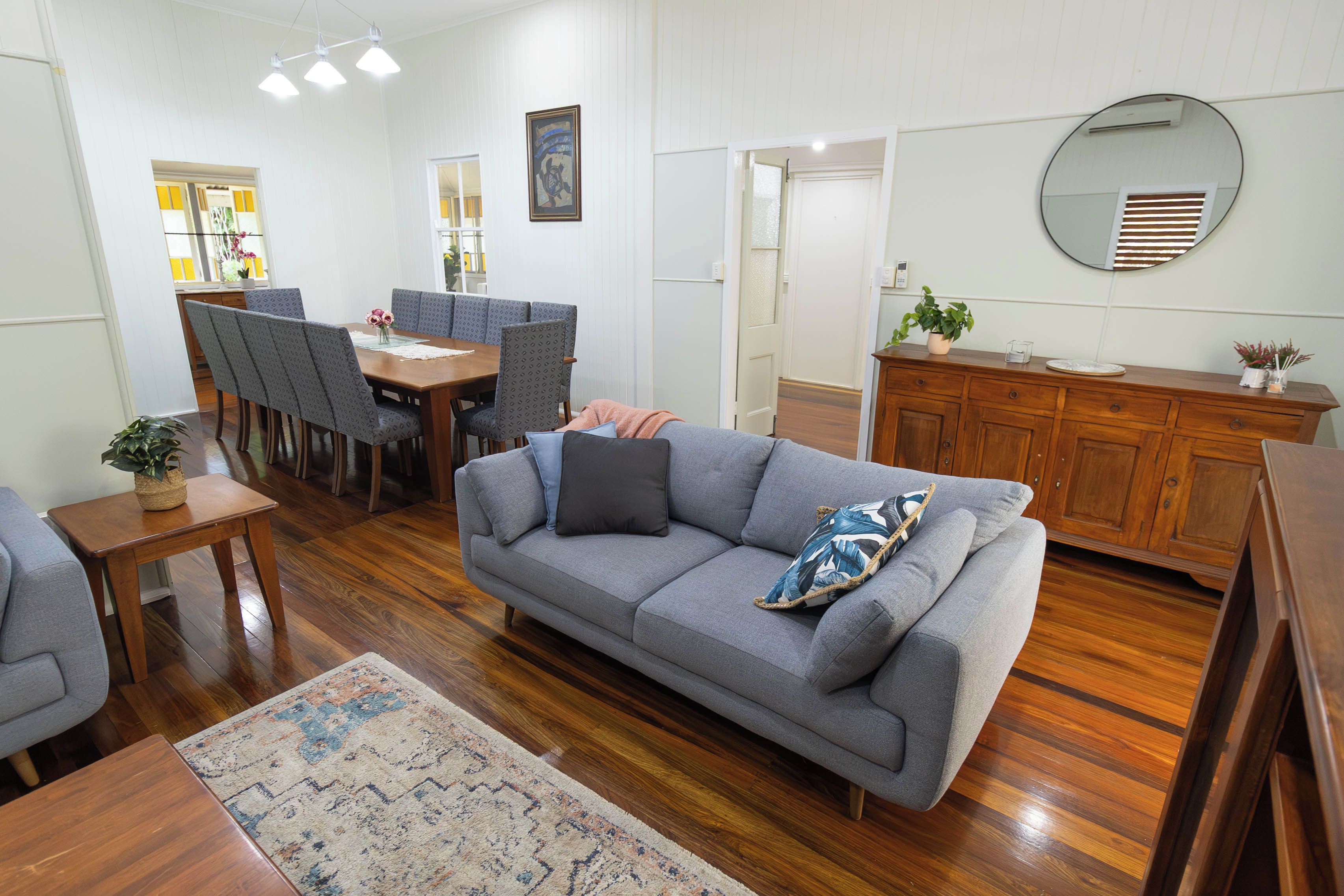 A living room with a grey sofa, wooden dining table, and chairs. There's a small table with a plant, a wooden cabinet, and a mirror on the wall.
