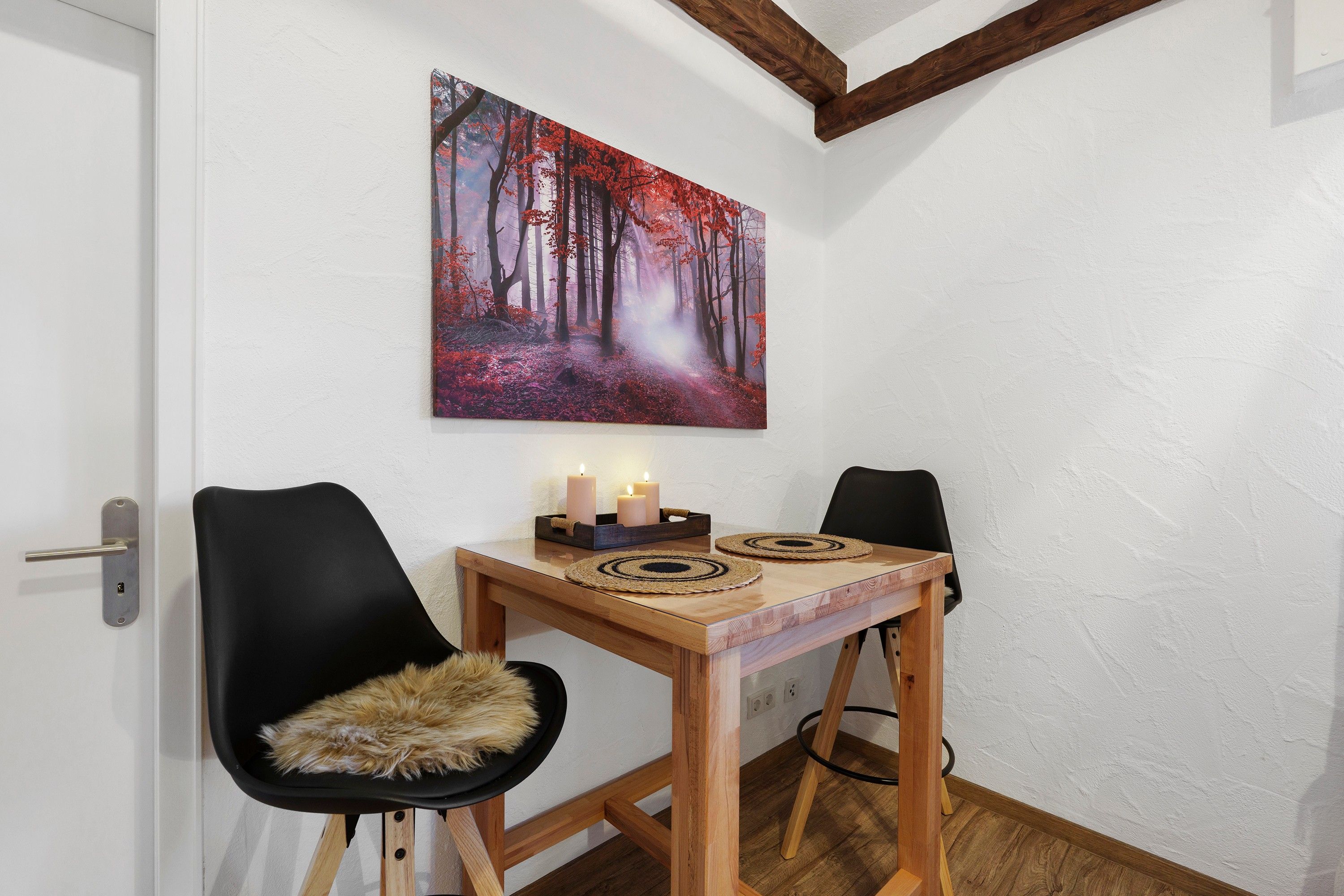 Un comedor con una mesa de madera, dos sillas, un cuadro de un bosque y velas sobre la mesa.