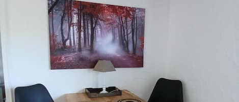 Et værelse med et træbord, to sorte stole, et billede af en skov og en lampe på bordet.