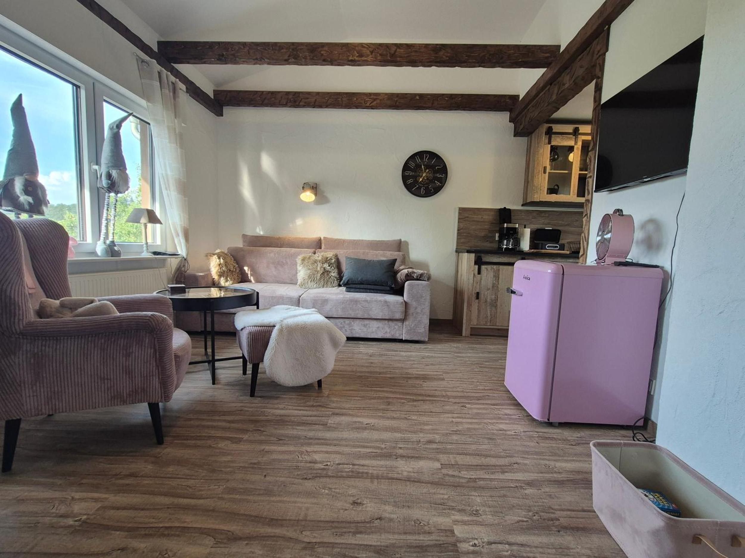 Ein gemütliches Wohnzimmer mit Sofa, Sesseln, Hocker, einem kleinen Tisch und einem pinken Kühlschrank.