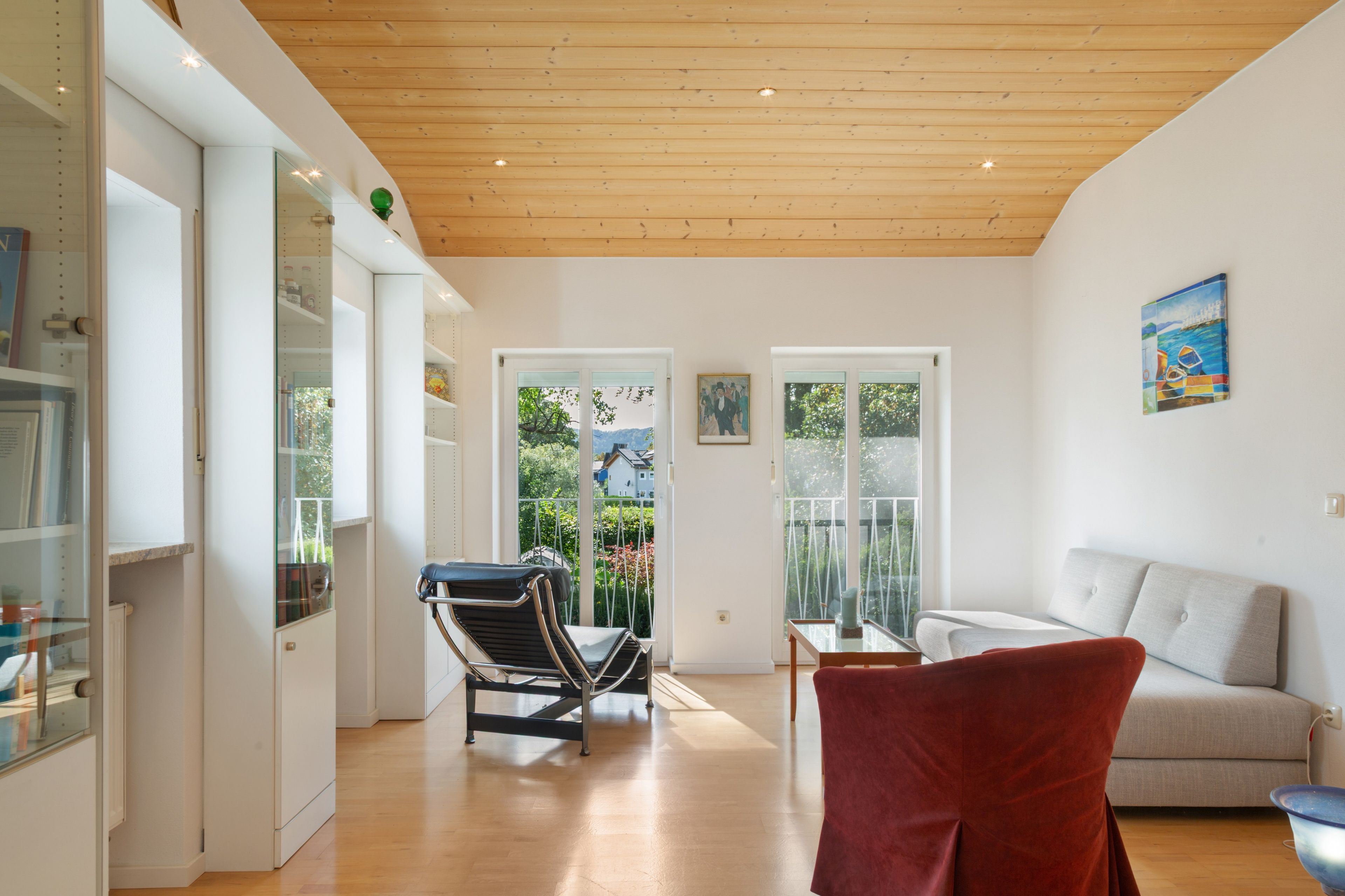 Ein modernes Interieur mit Holzbalkendecke, einem Essbereich mit Glastisch, einem roten Stuhl und einer Couch.