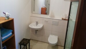 Casa de banho com lavatório branco, sanita e espelho sobre o lavatório. Há um banquinho pequeno e uma prateleira com toalhas.