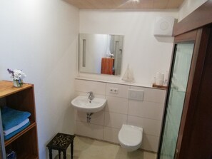 Ein Badezimmer mit einem weißen Waschbecken, einer Toilette und einem Spiegel über dem Waschbecken. Es gibt einen kleinen Hocker und ein Regal mit Handtüchern.