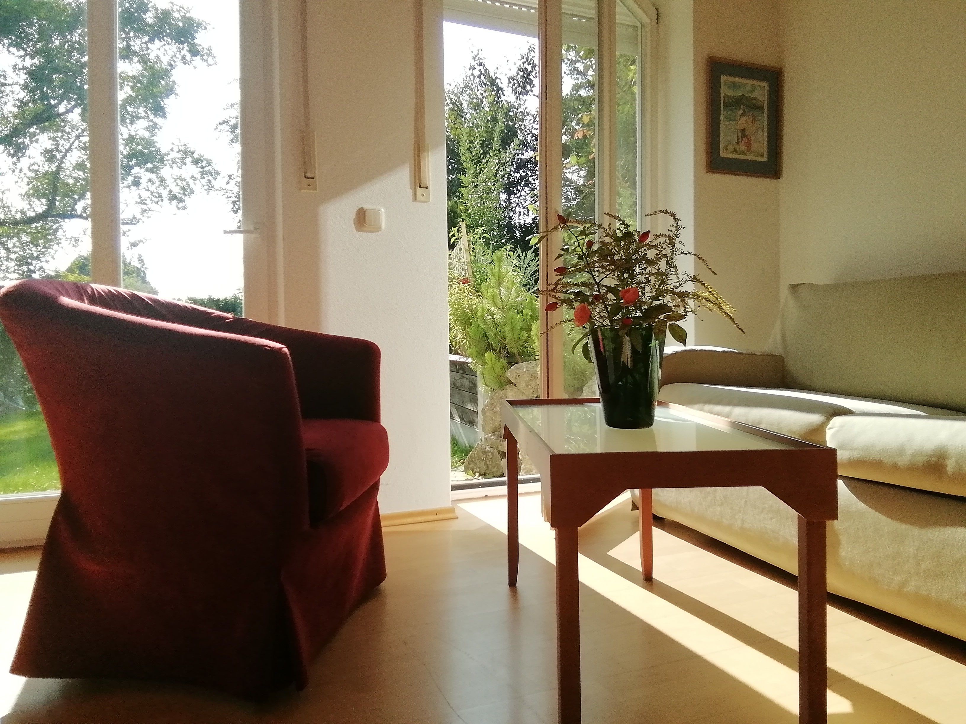 Ein Wohnzimmer mit einem roten Sessel, einem Holztisch, einem beigen Sofa und einem Fenster mit Blick auf Grünflächen.