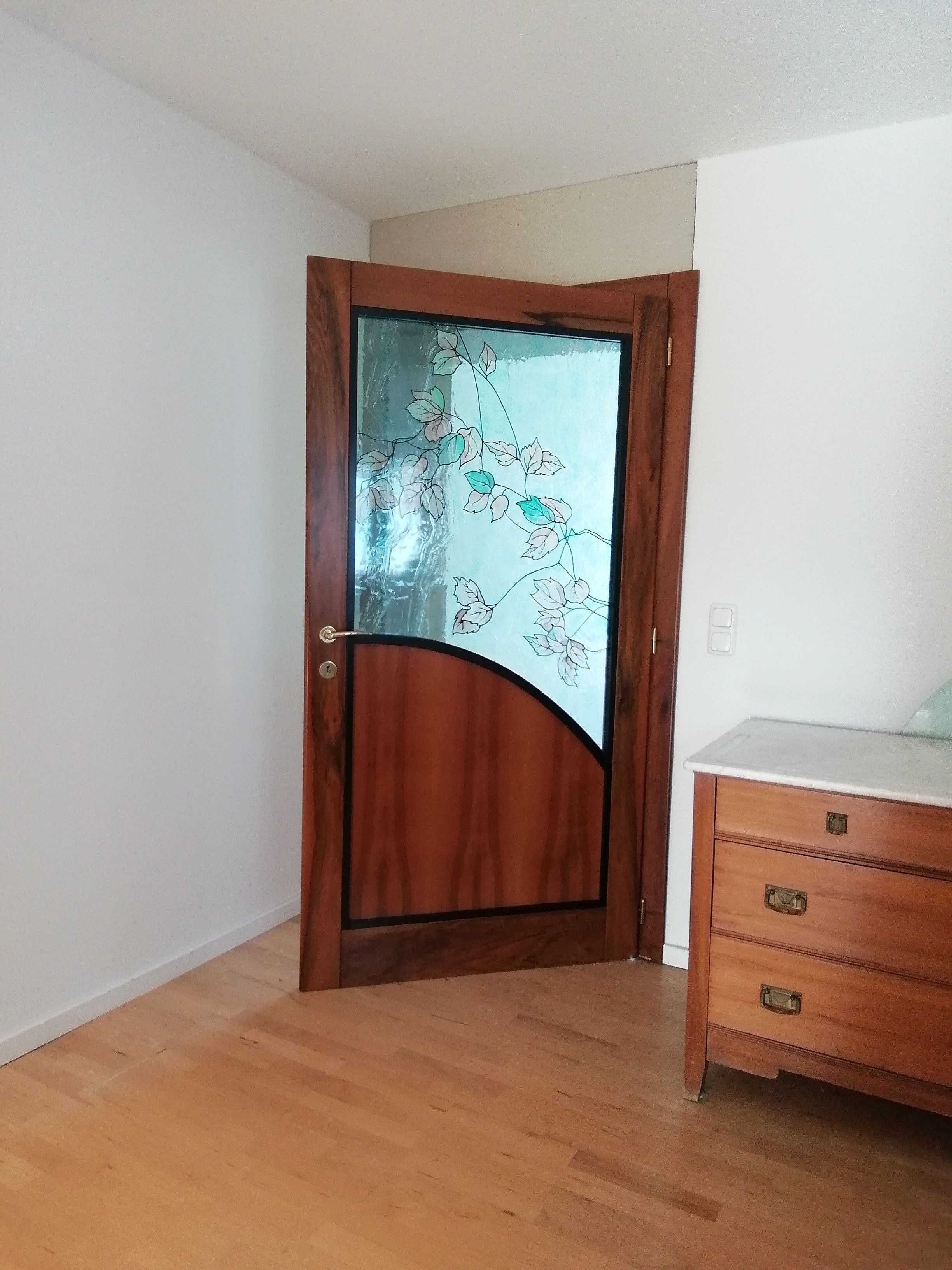 Ein Zimmer mit einer Holztür, die ein Fenster mit floralem Dekor enthält, einem Holzkommode mit Schubladen und einem hellen Boden.