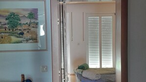 Um quarto com piso de madeira, um sofá azul, uma cama com cobertor xadrez, um quadro na parede e uma porta que dá para outro cômodo.