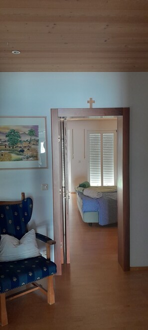 Ein Zimmer mit Holzboden, einem blauen Sofa, einem Bett mit kariertem Überwurf, einem Bild an der Wand und einer Tür zu einem anderen Raum.