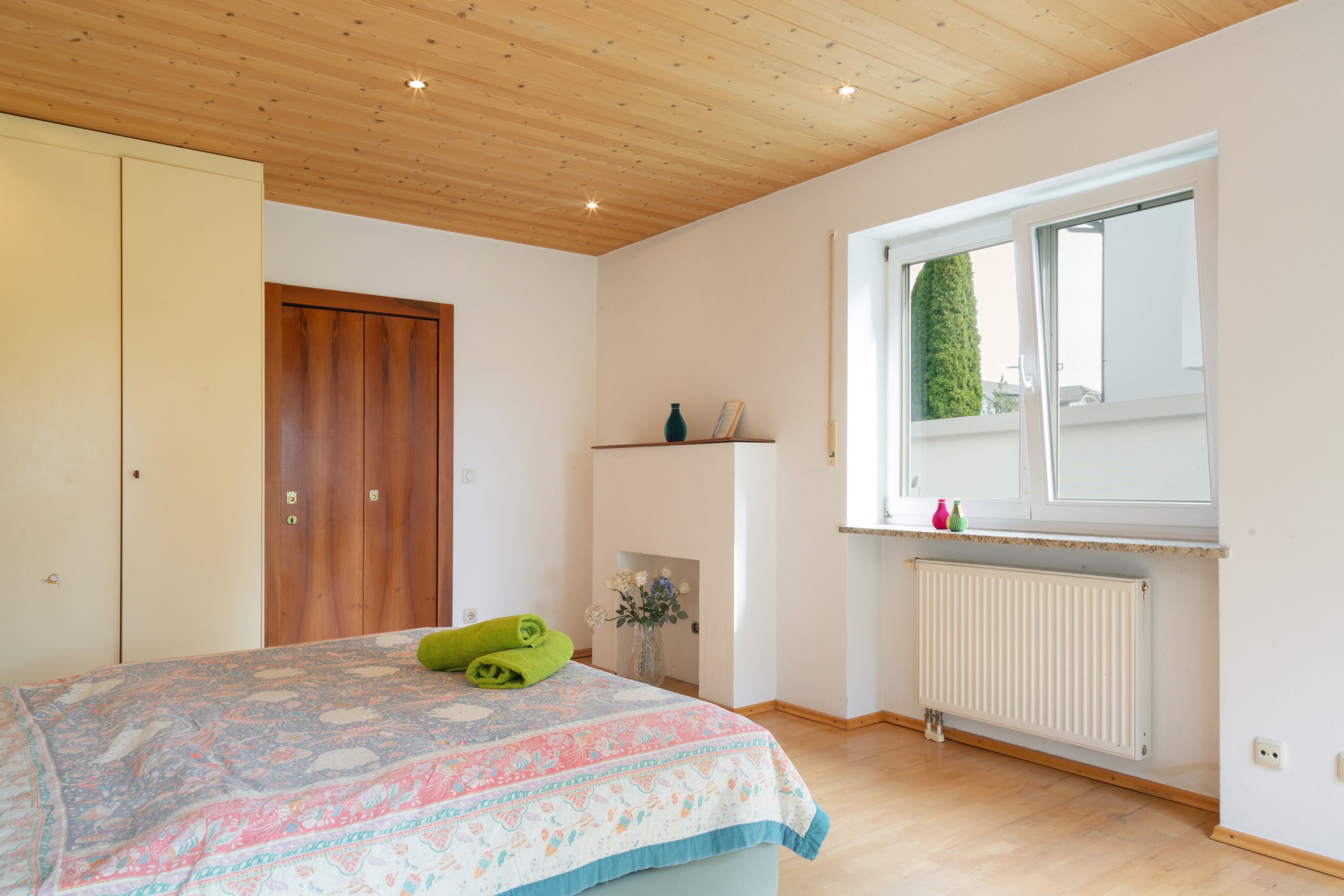 A bedroom with a wooden ceiling, a bed with a floral bedspread, a window with a view of greenery, and a radiator.