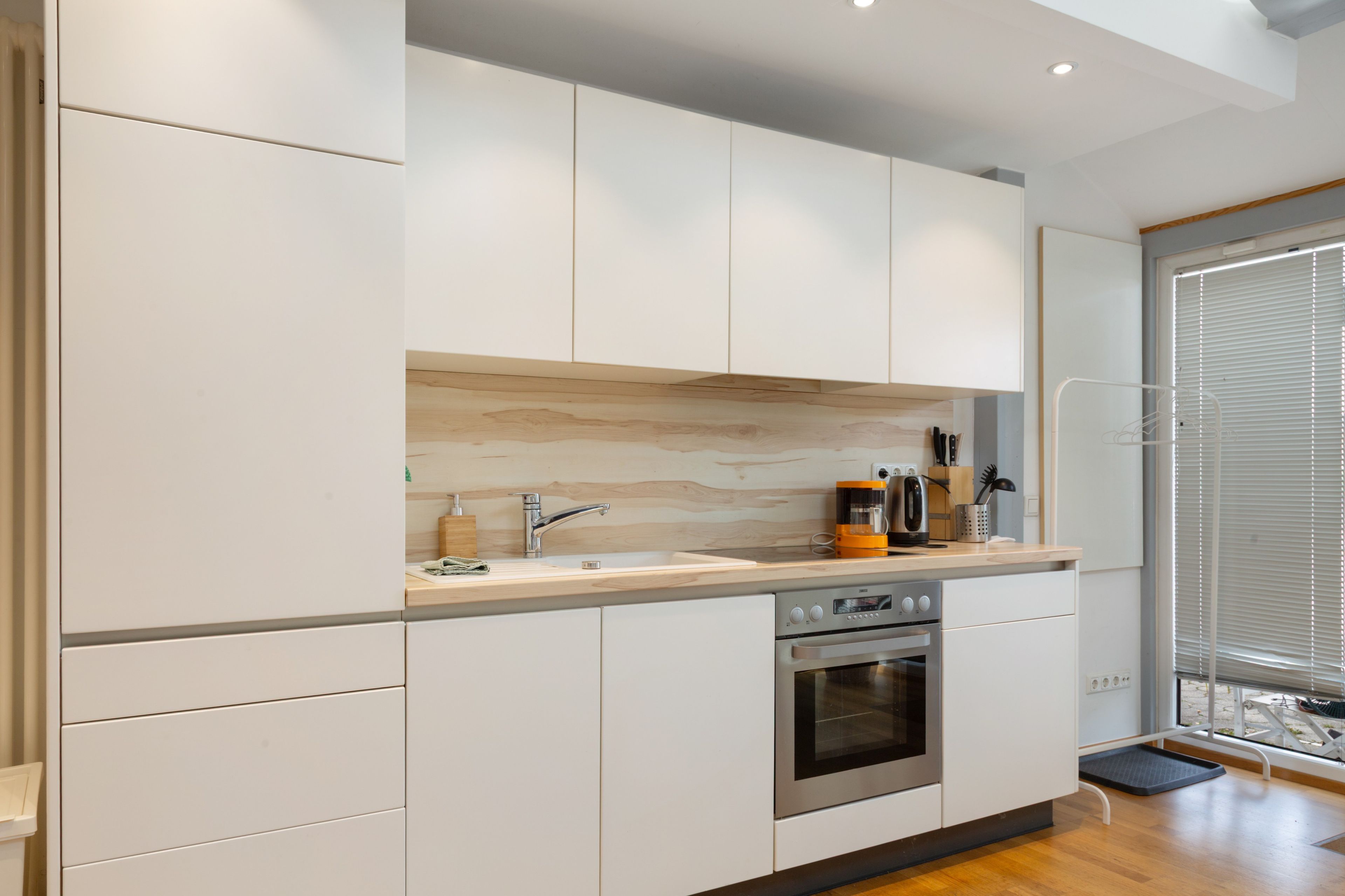 Een moderne keuken met witte kastjes, een houten aanrecht, een roestvrijstalen oven en een raam met zonwering.