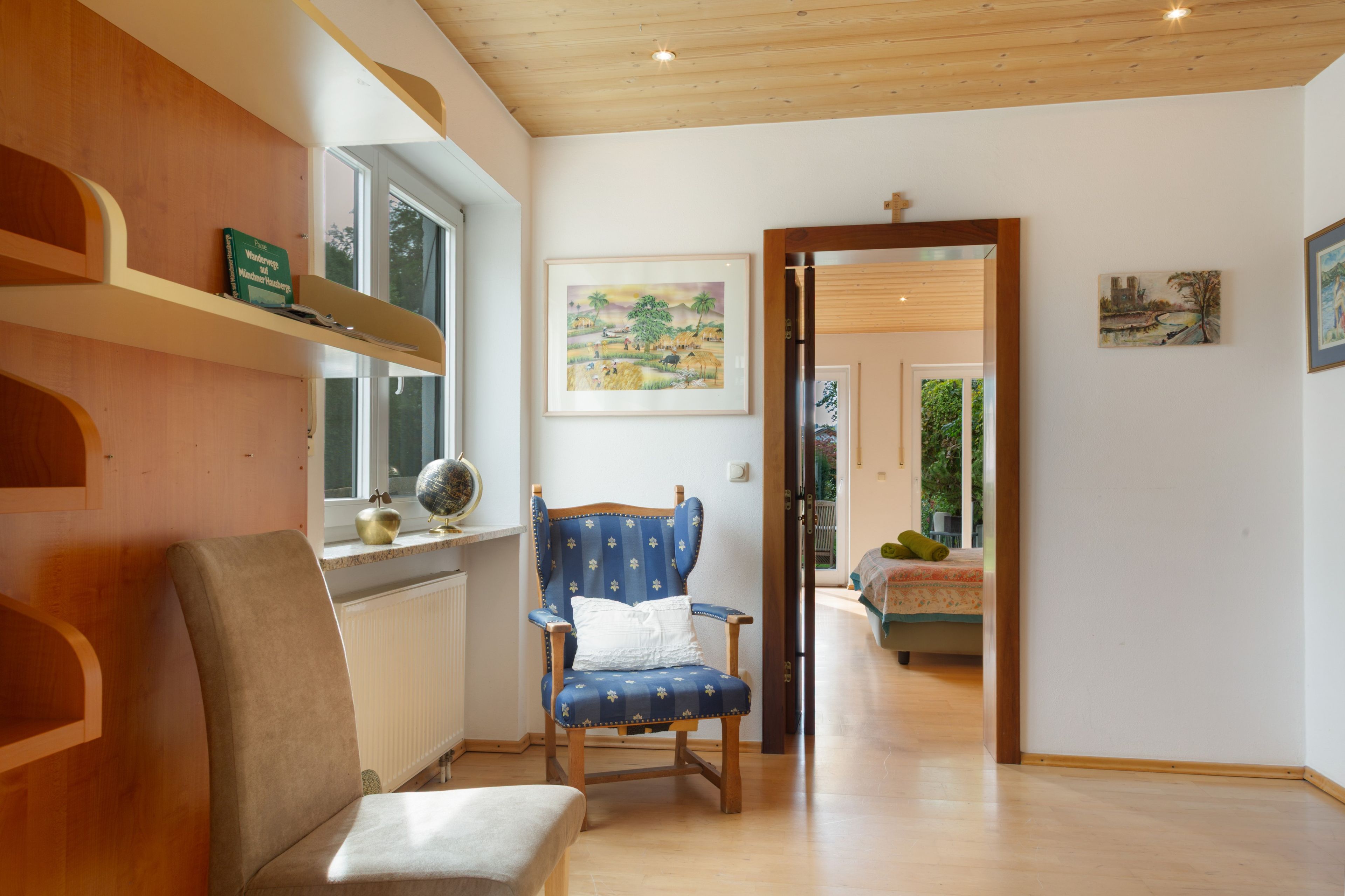 Une pièce avec un plancher en bois, un fauteuil recouvert de tissu bleu, un radiateur, une étagère remplie de livres et un tableau accroché au mur.