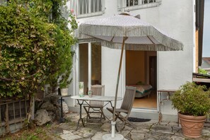 En udendørs terrasse med et bord og stole, en potteplante og en hvid bygning med et vindue.