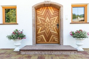 Eine Holztür mit geometrischem Muster, begleitet von zwei weißen Pflanzgefäßen mit Blumen, und einem Nummernschild darüber.