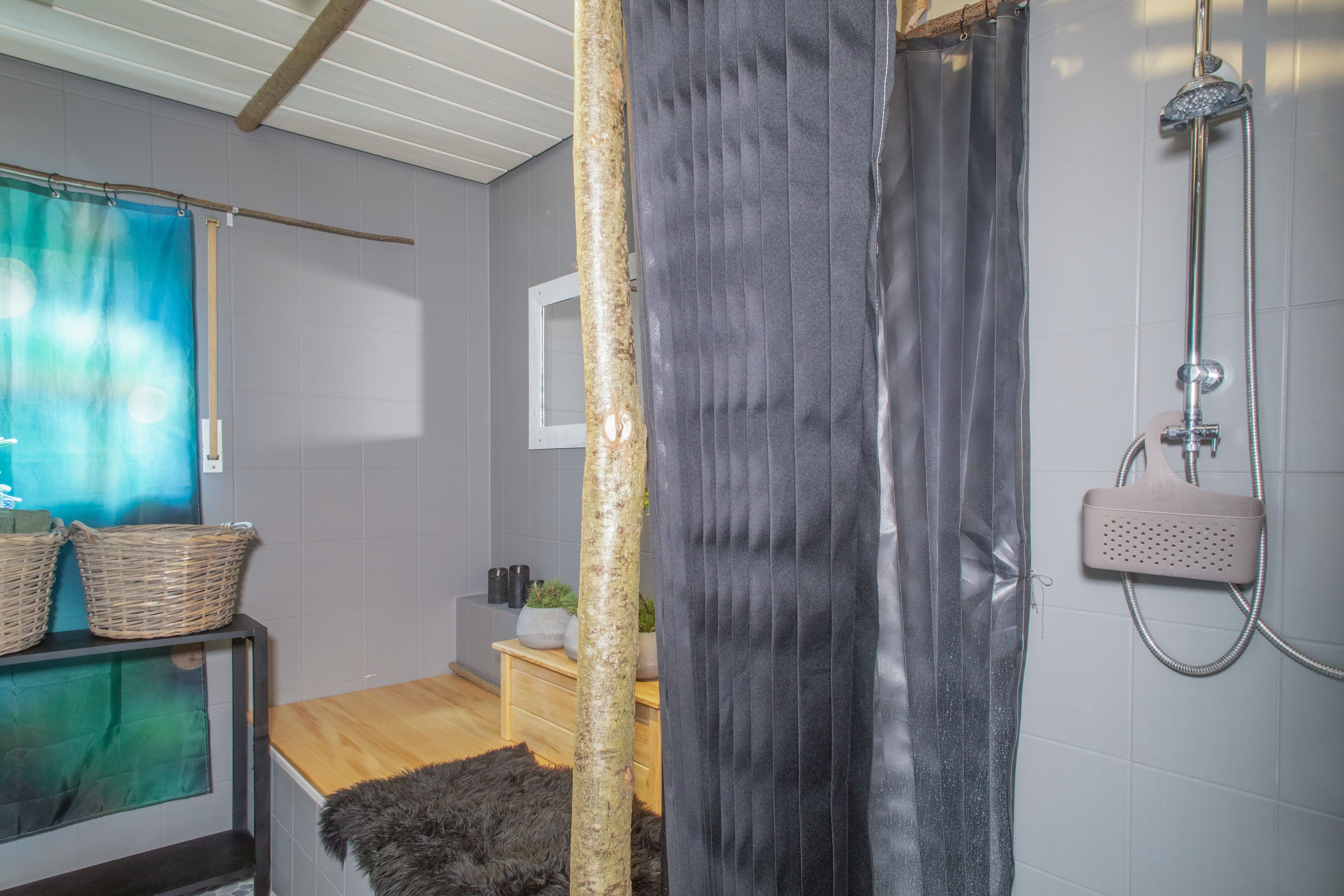 Ein Badezimmer mit Dusche, Holzboden, einem kleinen Tisch mit Körben und einem Fenster mit Vorhang.