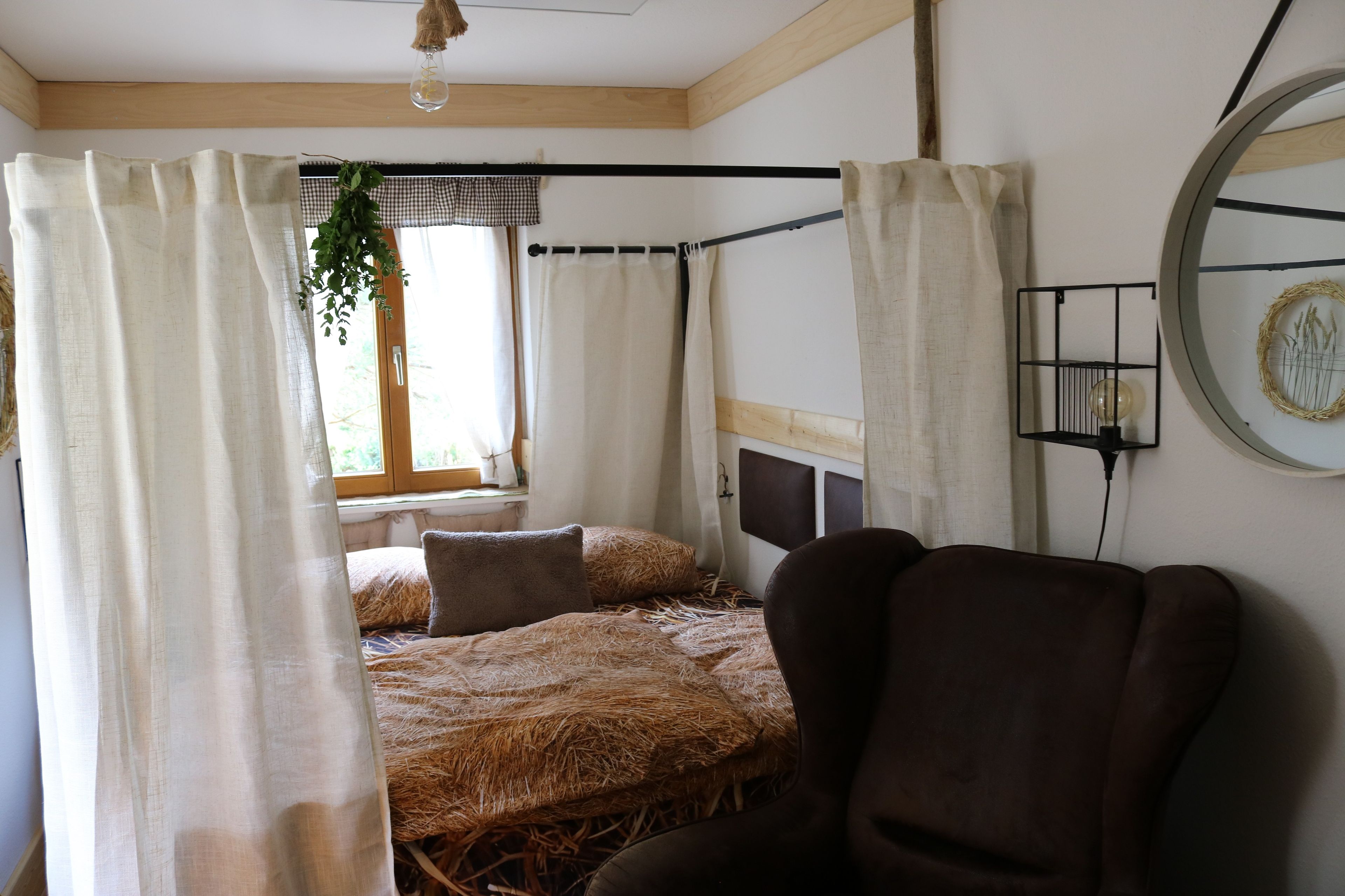Ein Zimmer mit einem strohbedeckten Bett, einem braunen Ledersessel und einem Fenster, an dem draußen eine Pflanze hängt.