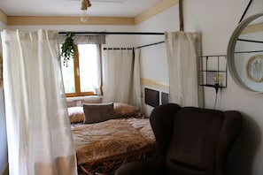 Una habitación con una cama cubierta de paja, un sillón de cuero marrón y una ventana con una planta colgando en el exterior.