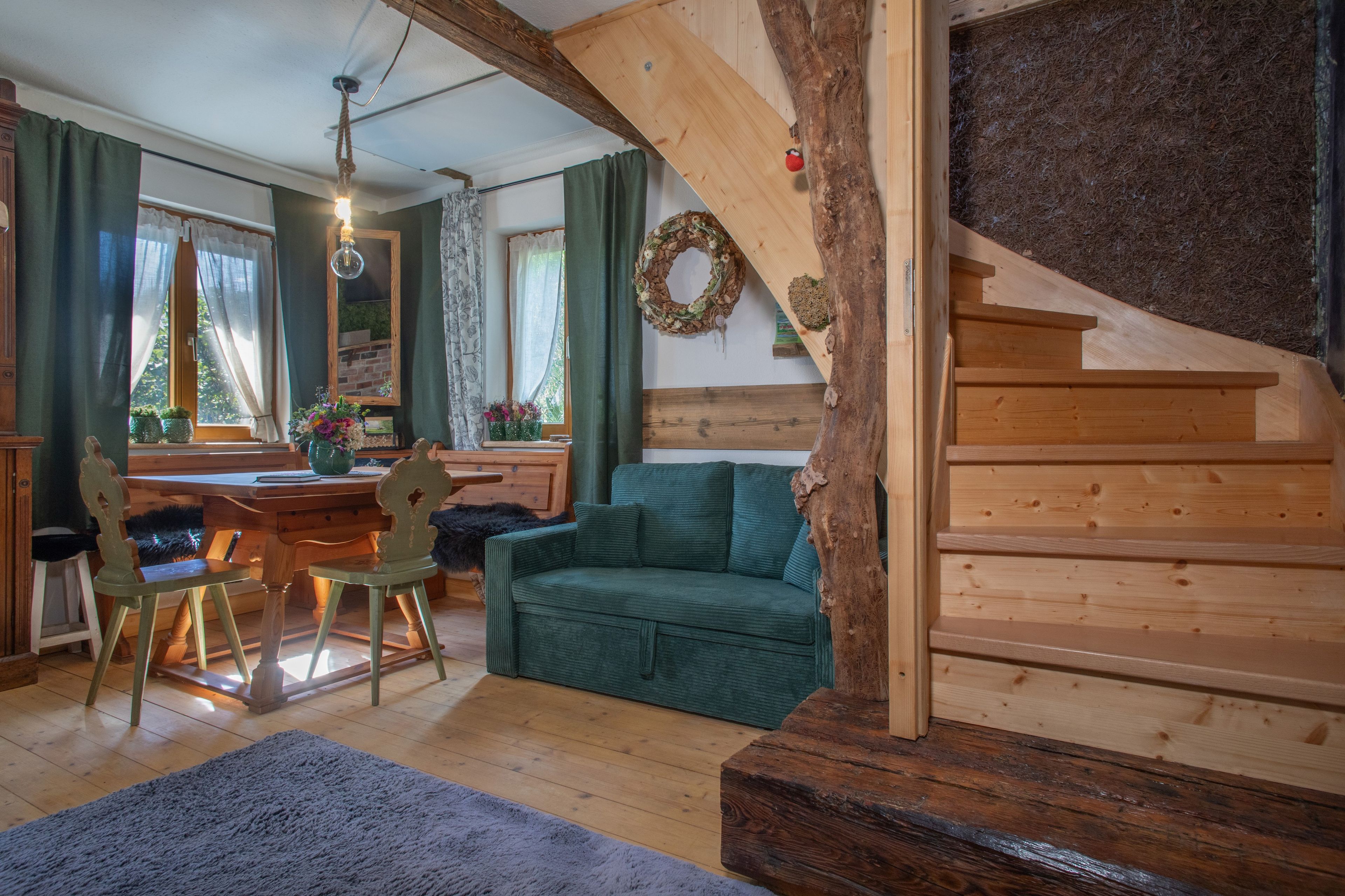 Uma sala de estar aconchegante com escada de madeira, área de jantar com mesa e cadeiras, e sofá verde.