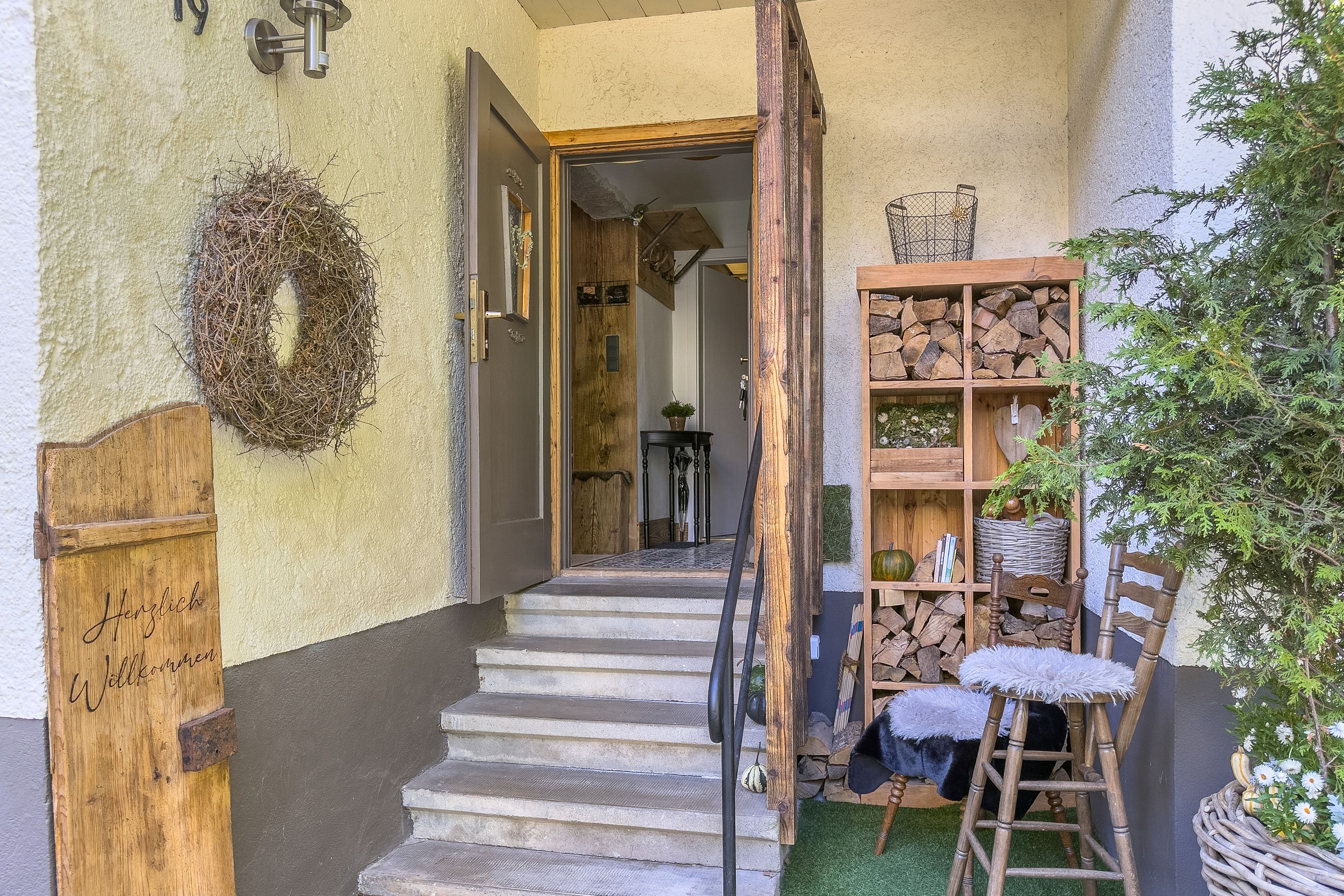 Eine Holztür mit einem Schild, eine Treppe, die zu einem Raum führt, ein Holzregal mit Holzscheiten und eine Pflanze.
