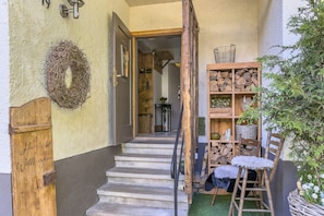 Eine Holztür mit einem Schild, eine Treppe, die zu einem Raum führt, ein Holzregal mit Holzscheiten und eine Pflanze.