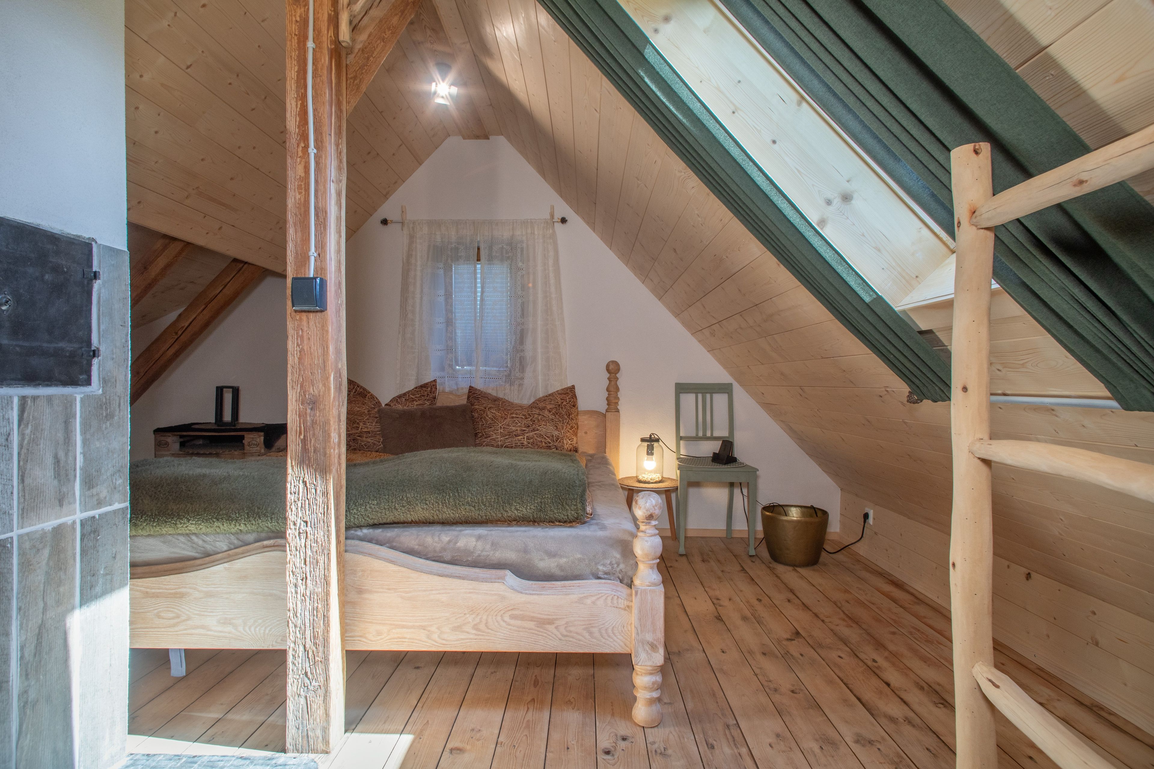Une chambre à coucher aménagée dans un espace mansardé, avec un plafond en pente, un lit en bois, une table de chevet, une lampe et une fenêtre ornée de rideaux légers.