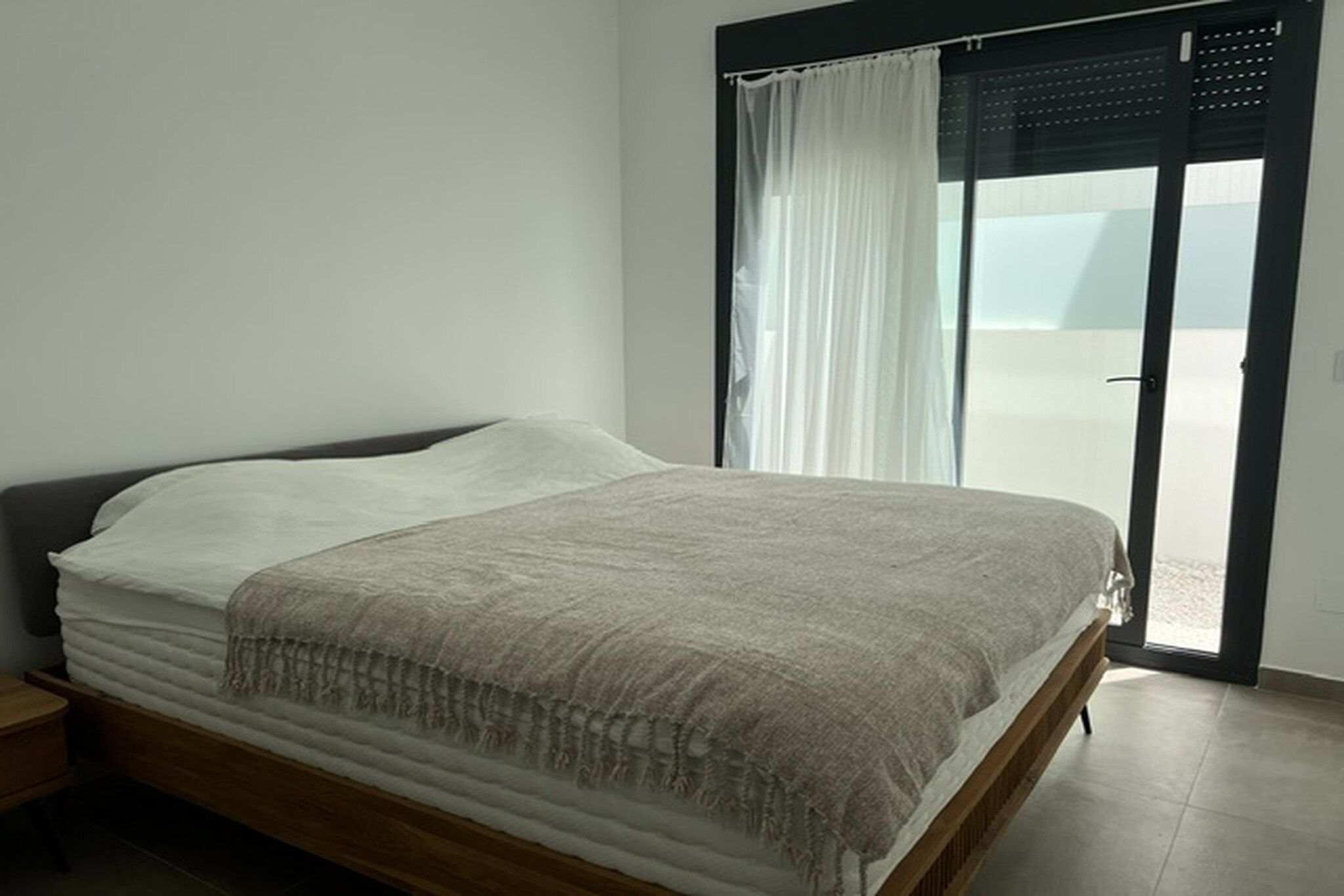 A single bed with a wooden frame, a white mattress, and a beige blanket. There is a window with white curtains and a view of the outside.