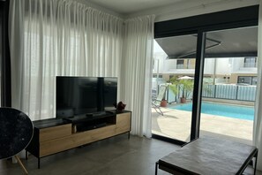 Ein modernes Wohnzimmer mit einem Flachbildfernseher auf einer Holzkonsole, einem Esstisch und Blick auf einen Swimmingpool durch eine Glasdoor.