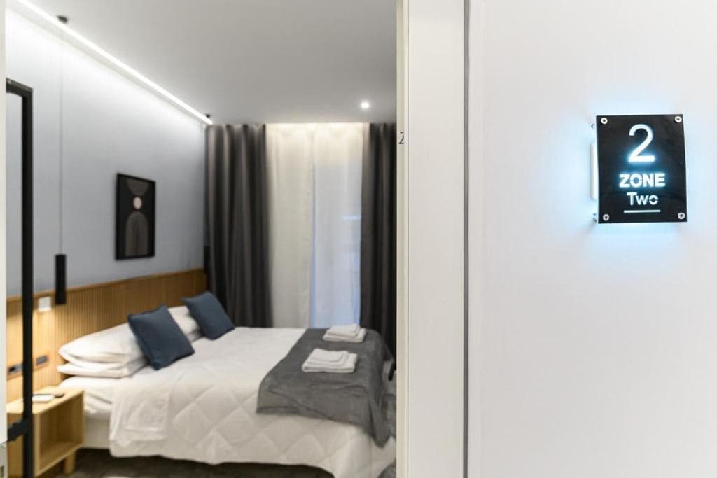 A hotel room with a bed, a nightstand, a wall-mounted sign indicating '2 Zone Two,' and a window with curtains.
