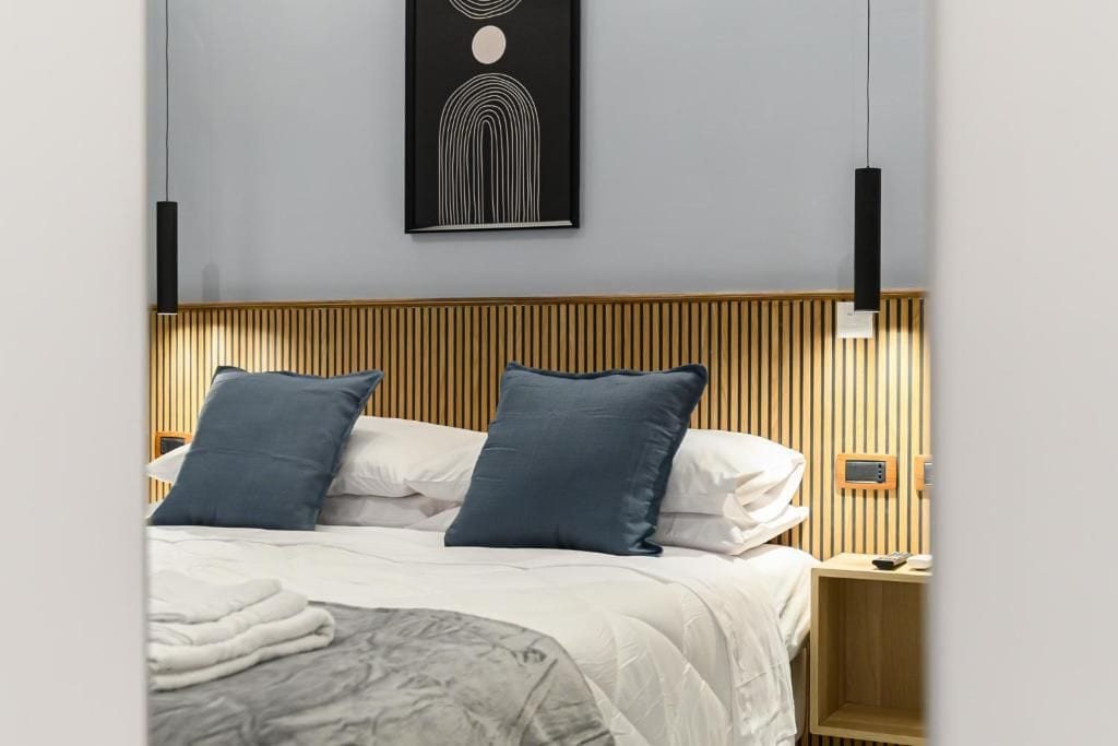 A modern bedroom with a wooden headboard, two pillows, a bedside table with a remote, and a wall-mounted artwork.