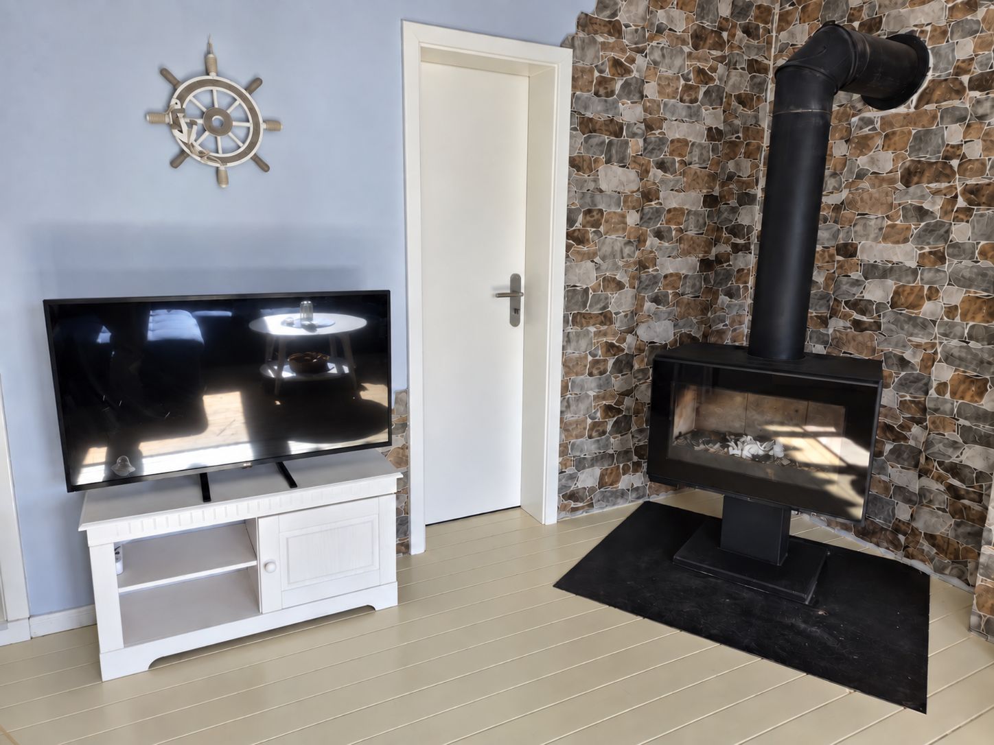 A living room with a television on a white stand, a stone wall with a fireplace, and a door.